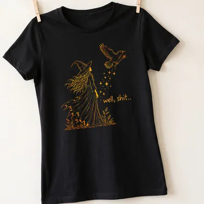 Line-art design featuring a witch and crow with the text 'well, shit..' in orange — printed on a women's shirt